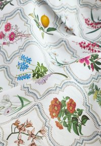 Tissu à motif floral avec des fleurs multicolores, des citrons jaunes et des feuilles sur fond blanc, bordé d'une couture décorative festonnée.