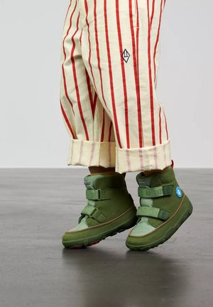 Child wearing beige pants with red stripes and green high-top shoes with two straps, standing on a gray floor.
