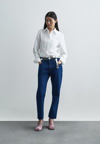 Λευκό πουκάμισο με μακριά μανίκια και κουμπιά μπροστά, συνδυασμένο με σκούρα μπλε τζιν παντελόνι. Το μοντέλο φοράει ανοιχτό ροζ πλατφόρμες. Λείες υφές, κομψή εφαρμογή.