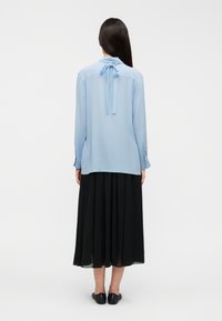 Ανοιχτό γαλάζιο μπλουζάκι με μακριά μανίκια και λεπτομέρεια δέσιμου στην πλάτη, συνδυασμένο με μαύρη πλισέ φούστα και μαύρα επίπεδα παπούτσια, εμφανίζεται από την πίσω πλευρά.