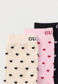 Trei perechi de șosete în culorile crem, roz și negru, cu mici modele de inimioare și numele parțial al brandului pe mansete, așezate suprapuse pe o suprafață albă.