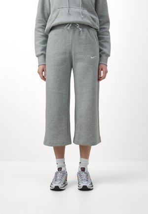 Persoon die een grijze cropped joggingbroek met Nike-logo, een grijze hoodie, witte Nike-sokken en witte sportsneakers draagt, staande op een witte vloer.