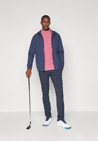 Veste à capuche navy sur un haut rose à demi-fermeture éclair, associée à un pantalon navy et à des chaussures de golf blanches avec des accents bleus, tenant un club de golf.