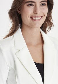 Blazer bianco con revers a punta, realizzato in tessuto liscio, indossato sopra una maglietta nera. Il tono della pelle è visibile nell'area del collo e del petto.