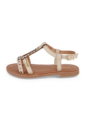 Sandale en cuir beige avec design découpé et accent doré. Bride de cheville ajustable et semelle plate. Texture lisse avec détails couturés.