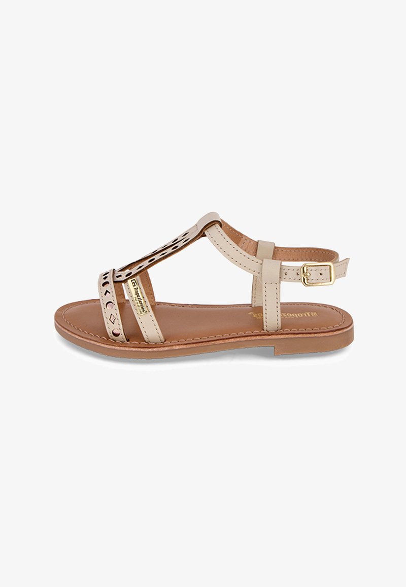 Sandale en cuir beige avec design découpé et accent doré. Bride de cheville ajustable et semelle plate. Texture lisse avec détails couturés.