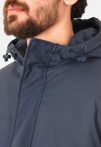 Giacca a vento blu navy con cappuccio, caratterizzata da un colletto alto, chiusura con zip, cordino regolabile e tessuto liscio e resistente all'acqua.