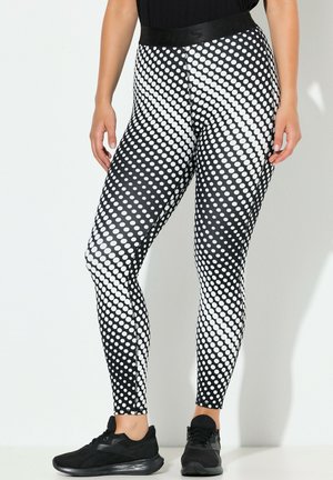 Vrouw die zwarte en witte leggings met polkadots en zwarte sportschoenen draagt, staand tegen een lichtgrijze muur.