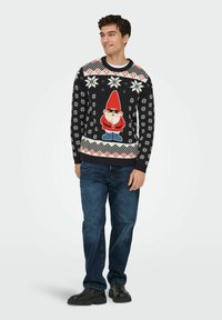 Joven vestido con un suéter festivo negro que tiene un gnomo con gafas de sol y patrones de copos de nieve blancos, jeans oscuros y botas negras.