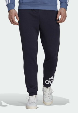 Homme portant un pantalon de survêtement bleu marine avec le logo Adidas blanc sur la jambe et des baskets blanches, marchant sur un fond clair.