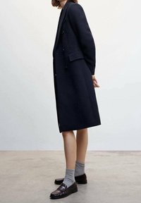 Cappotto lungo in lana navy con design doppiopetto, spacchi laterali e spalle strutturate, abbinato a mocassini scuri e calzini grigi.