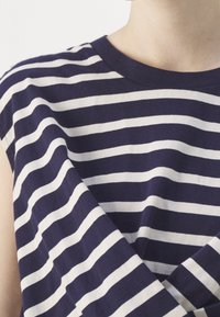 Personne portant un t-shirt rayé horizontal bleu marine et blanc, bras croisés, montrée du cou jusqu’au milieu du torse sur fond blanc.