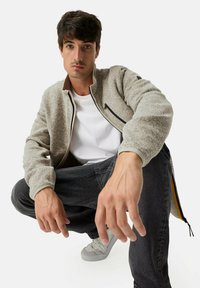 Grau strukturierte Jacke mit hohem Kragen, Reißverschluss-Taschen vorne, beigen Akzenten, kombiniert mit schwarzen Jeans und grauen Sneakers.