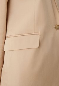 Beige blazertas met overslag, close-up van de gestructureerde stof en een enkele tan knop zichtbaar aan de rechterkant.