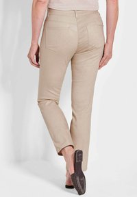 Beige, taillierte Hose mit glatter Textur, die subtile Muster und Gesäßtaschen aufweist, kombiniert mit einer schwarzen Riemensandale.