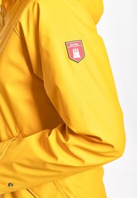 Giacca impermeabile gialla con una texture liscia, caratterizzata da una toppa a forma di scudo rossa sul maniche e un design con colletto alto.