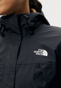 Gros plan d'une personne portant une veste imperméable noire North Face avec une capuche ajustable et un logo blanc sur la poitrine.