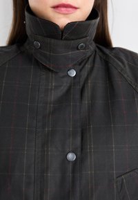 Veste à carreaux noire avec un col montant, présentant des boutons-pression noirs et une poche. Le matériau semble lisse avec une texture subtile.