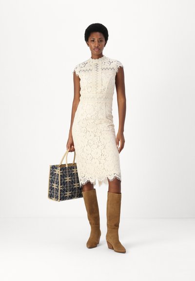 Robe en dentelle couleur crème, ajustée avec un ourlet festonné et un col haut ; portée avec des bottes en daim beige jusqu'aux genoux et un sac fourre-tout à motifs bleu marine.