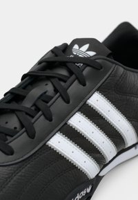 Black Adidas sneaker with white three stripes, black laces, and visible Adidas logo on tongue and sole side.