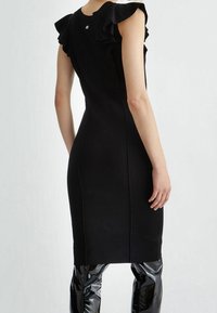 Robe noire ajustée avec des accents de volants aux épaules, un corsage texturé et un détail subtil de boutons à l'arrière. Portée avec un pantalon noir brillant.