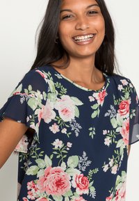 Blusa floral azul-marinho de mangas curtas. Apresenta padrões de rosas em rosa e verde, um decote redondo e uma textura leve e semi-transparente.