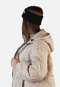 Fascia per capelli nera a coste indossata con un giubbotto trapuntato color crema, dotato di cappuccio e chiusura con zip. La fascia presenta un piccolo etichetta sul lato.