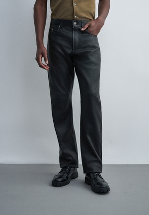 Mees kannab musti sirgete jalgadega nahkpükse, musti paeltega kingi ja pruuni särki, seisab ühevärvilise tausta ees.