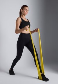 Svart idretts-bh og høyt midje leggings, med et gult motstandsbånd i bruk. Modellen står på svarte sneakers og viser frem treningsklær.