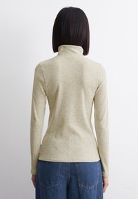 Vrouw met steil, donker haar, gekleed in een lichtbeige gemêleerde coltrui met lange mouwen en blauwe spijkerbroek, met de rug naar een witte muur gekeerd.