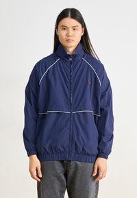 Marineblå vindjakke med hvid piping, lynlås foran, høj krave og elastiske manchetter; båret over grå jeans.