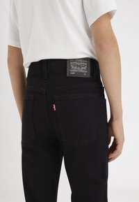 Jeans de mezclilla negra con un ajuste de cintura alta, que cuentan con bolsillos traseros, un logo en forma de etiqueta roja y una etiqueta de marca gris. La tela tiene una textura suave.