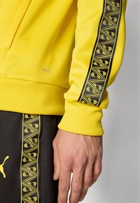 Gul sweatshirt med ribbede manchetter, der har en sort stribe med et geometrisk gult mønster og et lille brandinglabel.
