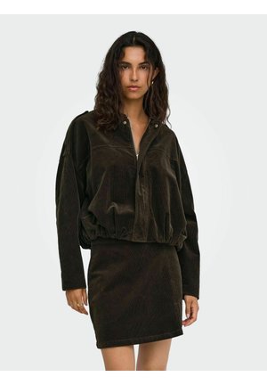 Femme aux cheveux bruns ondulés portant une veste zippée en velours côtelé marron foncé et une jupe assortie sur un fond clair uni.