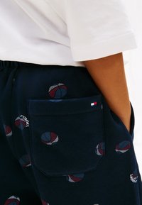 Παιδί που φοράει ναυτικό μπλε φόρμες με εκτυπώσεις καλαθοσφαίρισης και λογότυπο Tommy, καθώς και μια μικρή σημαία στην πίσω τσέπη, συνδυασμένα με λευκό μπλουζάκι.