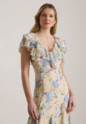 Γυναίκα με ανοιχτά καστανά μαλλιά, ντυμένη με παστέλ floral φόρεμα με βολάν στο V-λαιμό και κοντά μανίκια, μπροστά σε απλό ουδέτερο φόντο.