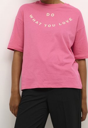 Osoba nosząca luźny różowy t-shirt z białym napisem "RÓB TO, CO KOCHASZ" oraz czarne spodnie na jednolitym, jasnym tle.