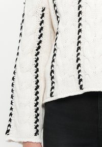 Chunky witte gebreide trui met zwarte gevlochten accenten langs de naden en een cropped ontwerp. Textuur van kabelpatroon zichtbaar.