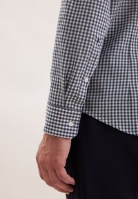 Braccio maschile che indossa una camicia a maniche lunghe a scacchi bianco e nero con polsini abbottonati di bianco, poggiato accanto a pantaloni neri.