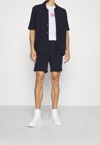 Camisa de manga corta azul marina, camiseta gráfica blanca debajo, pantalones cortos a juego y zapatillas altas blancas. Diseño simple con un corte casual.
