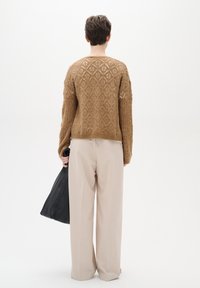 Gebreide trui in lichtbruin met diamantpatronen, gedragen bij crèmekleurige brede broekspijpen. Model draagt een zwarte leren tas.