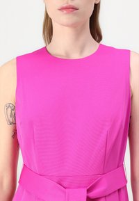 Přiléhavé šaty v živé růžové barvě, s kulatým výstřihem, bez rukávů a s opaskem v pase pro zvýraznění detailu.