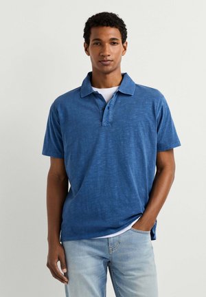 Joven con una camisa azul de manga corta con cuello y vaqueros azul claro, de pie con una mano en el bolsillo sobre un fondo liso.