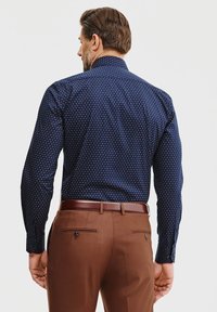 Granatowa koszula w mały wzór w białe i czerwone kropki; długie rękawy, mankiety na guziki; zestawiona z brązowymi spodniami; wyraźny kołnierz.