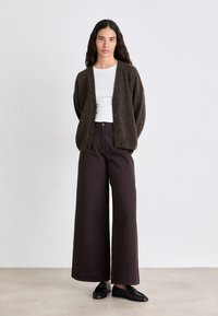 Cardigan lavorato a maglia marrone scuro con taglio loose, caratterizzato da un collo a V e bottoni frontali, abbinato a pantaloni larghi e a vita alta di colore scuro. Mocassini neri.