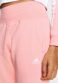 Vaatekankaalta pehmeät vaaleanpunaiset collegehousut, joissa on korkea vyötärö, sivutaskut ja valkoinen Adidas-logo vasemmassa lahkeessa. Sileä pinta.