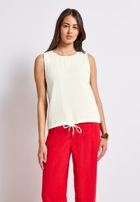Ljus beige ärmlös topp med en strukturerad yta och rund halsringning, med en dragsko i nederkant. Kombinerad med lysande röda linnebyxor.