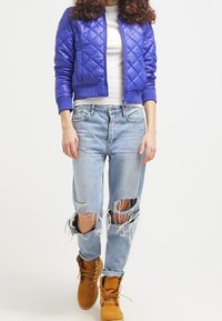 Giacca bomber trapuntata viola, top bianco aderente, jeans chiari sdruciti con strappi e stivali beige con lacci.