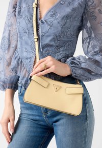 Borsa gialla con texture e logo triangolare, dotata di due cinturini piatti e design sottile. Abbinata a una blusa blu a fiori e jeans in denim.