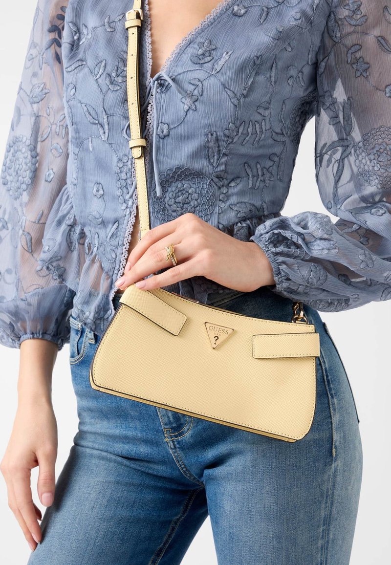 Borsa gialla con texture e logo triangolare, dotata di due cinturini piatti e design sottile. Abbinata a una blusa blu a fiori e jeans in denim.